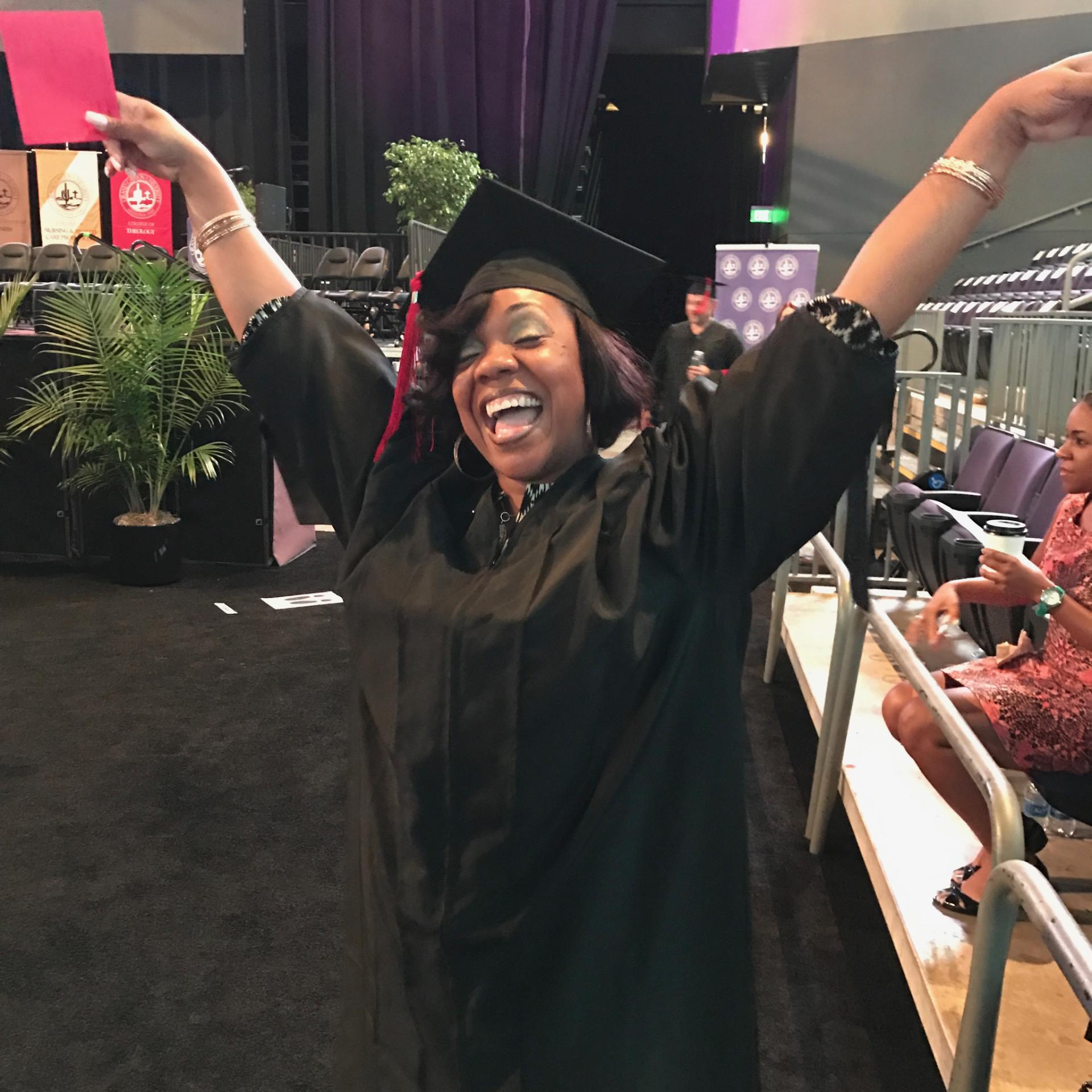 Marsha celebrating with arms raised in cap and gown at her seminary graduation.
