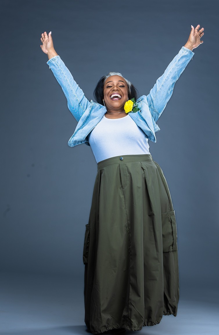 Marsha with both arms raised in joy, wearing a denim jacket and olive skirt.